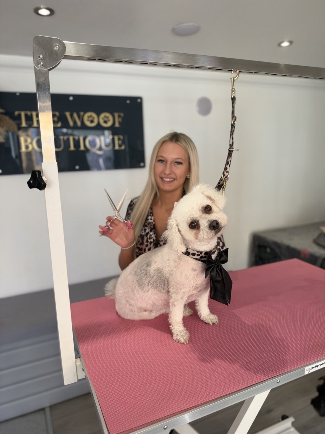 Charlotte smiling with a freshly groomed white fluffy dog on the pink grooming table at The Woof Boutique